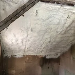 Poddasze z widoczną warstwą białej pianki izolacyjnej, przytwierdzonej metalowymi elementami, w trakcie prac ociepleniowych; widoczne drewniane elementy konstrukcyjne i fragment ściany