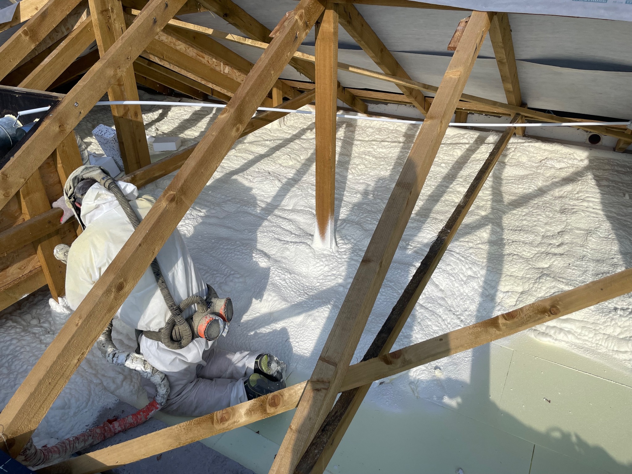 Pracownik w kombinezonie natryskuje białą pianę izolacyjną PUR na drewnianą konstrukcję dachu poddasza, tworząc jednolitą warstwę ocieplenia.