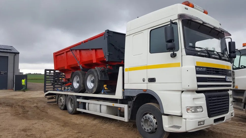 Ciężarówka DAF XF z czerwoną naczepą wywrotką na platformie transportowej, widoczna osoba w odblaskowej kamizelce i budynek w tle.