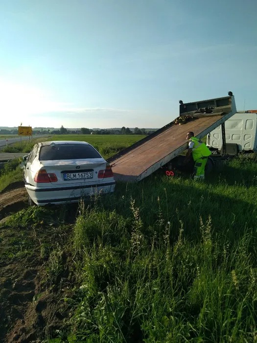 Biały samochód osobowy z uszkodzonym tyłem, z numerami rejestracyjnymi BLM 16753, wciągany na lawetę pomocy drogowej na tle zieleni i błękitnego nieba.