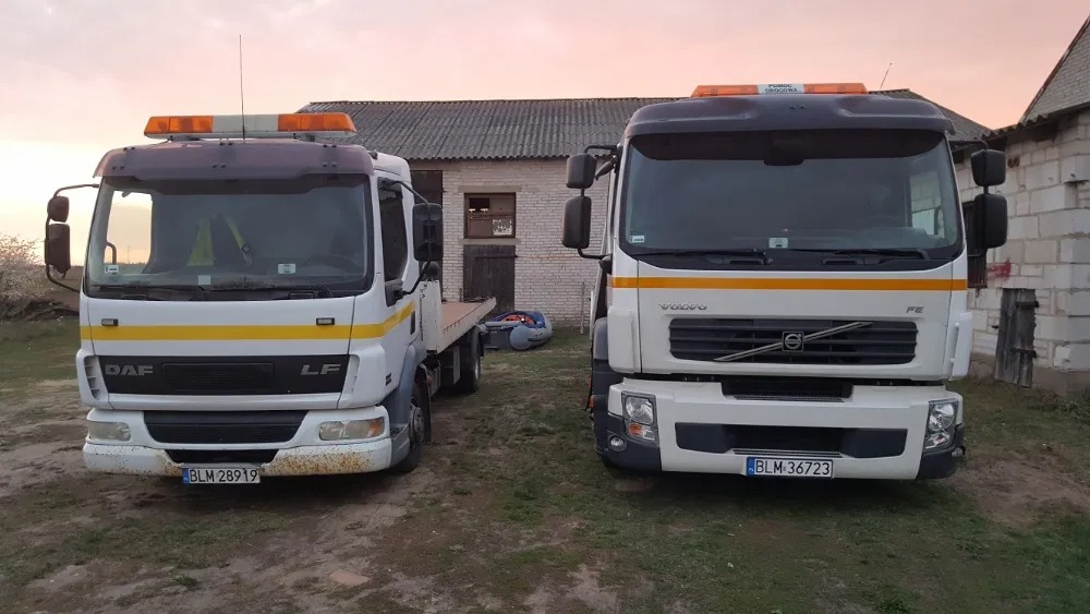 Dwa samochody ciężarowe: biały DAF LF z platformą i biały Volvo FE z pomarańczowymi kogutami na dachu, zaparkowane na trawie przed budynkiem gospodarczym.