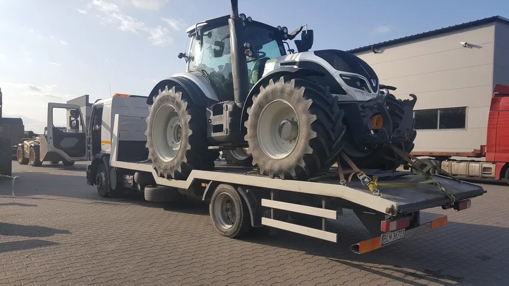 Biały ciągnik rolniczy zabezpieczony pasami na niskopodwoziowej platformie transportowej, widoczny numer rejestracyjny pojazdu.