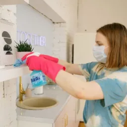 Osoba w masce i różowych rękawicach czyści półkę nad umywalką w jasnym wnętrzu, używając niebieskiego sprayu i ściereczki.