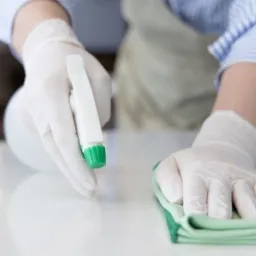 Osoba w rękawicach ochronnych czyści biały blat za pomocą zielonej ściereczki i spryskiwacza z zieloną końcówką.