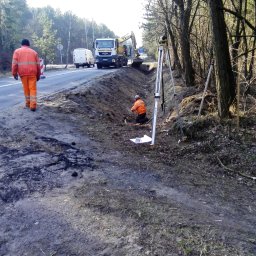 MS ROADWAY SPÓŁKA Z OGRANICZONĄ ODPOWIEDZIALNOŚCIĄ - Pracownik w pomarańczowym kombinezonie idzie poboczem drogi, w tle widoczny pracownik mierzący teren przy użyciu niwelatora, koparka przy wykopie i ciężarówka MAN