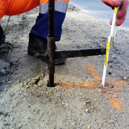 MS ROADWAY SPÓŁKA Z OGRANICZONĄ ODPOWIEDZIALNOŚCIĄ - Pomiar głębokości otworu w ziemi przy pomocy metalowego pręta i rozkładanej miarki, oznaczenia pomarańczową farbą na ziemi, pracownik w odzieży roboczej.