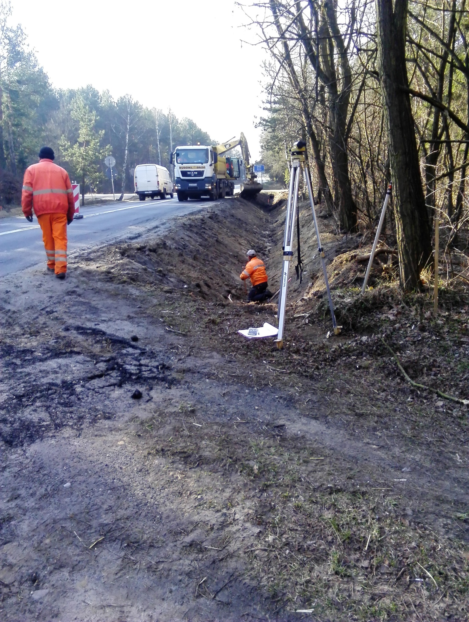 Pracownik w pomarańczowym kombinezonie idzie poboczem drogi, w tle widoczny pracownik mierzący teren przy użyciu niwelatora, koparka przy wykopie i ciężarówka MAN