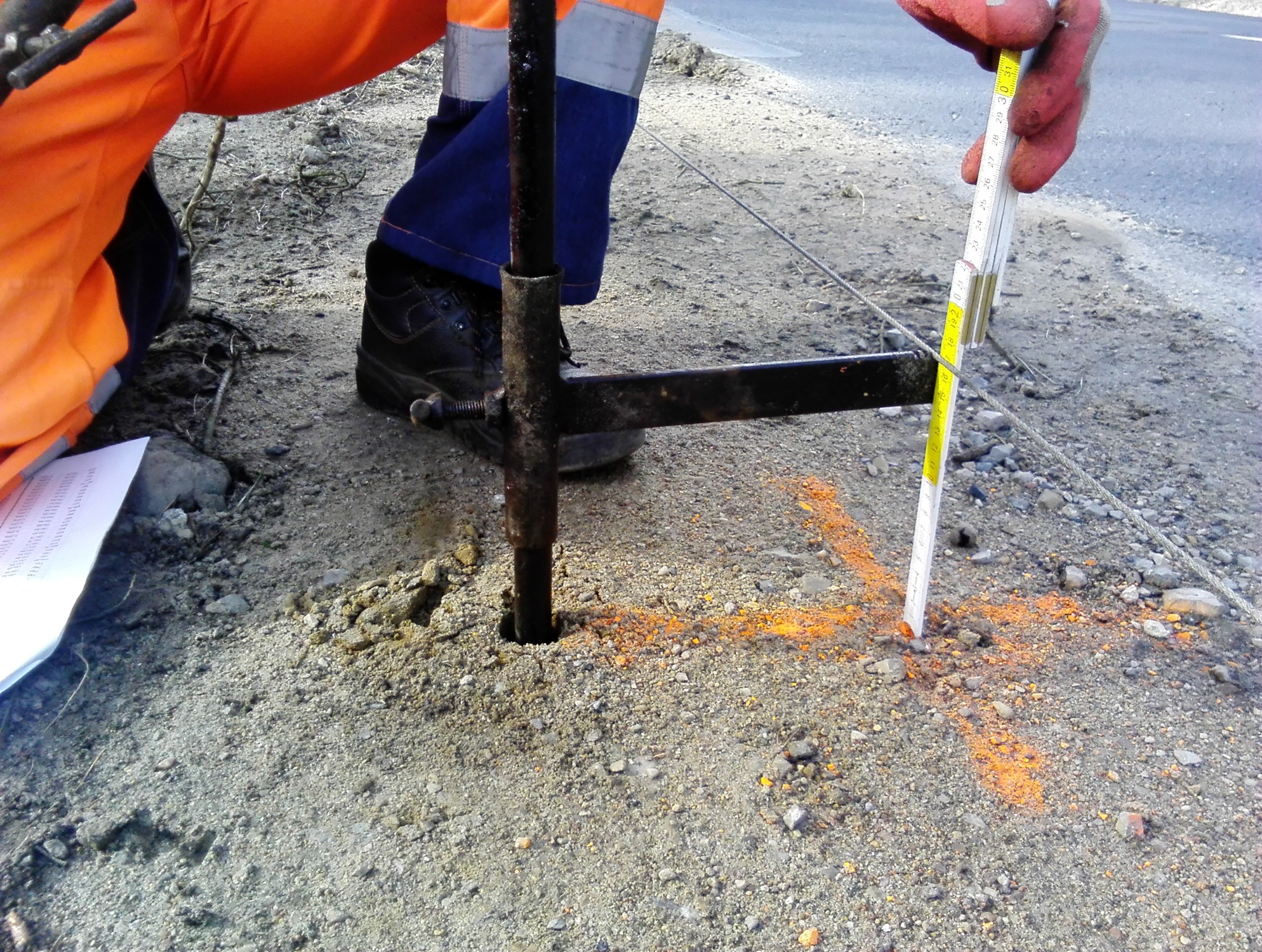 Pomiar głębokości otworu w ziemi przy pomocy metalowego pręta i rozkładanej miarki, oznaczenia pomarańczową farbą na ziemi, pracownik w odzieży roboczej.