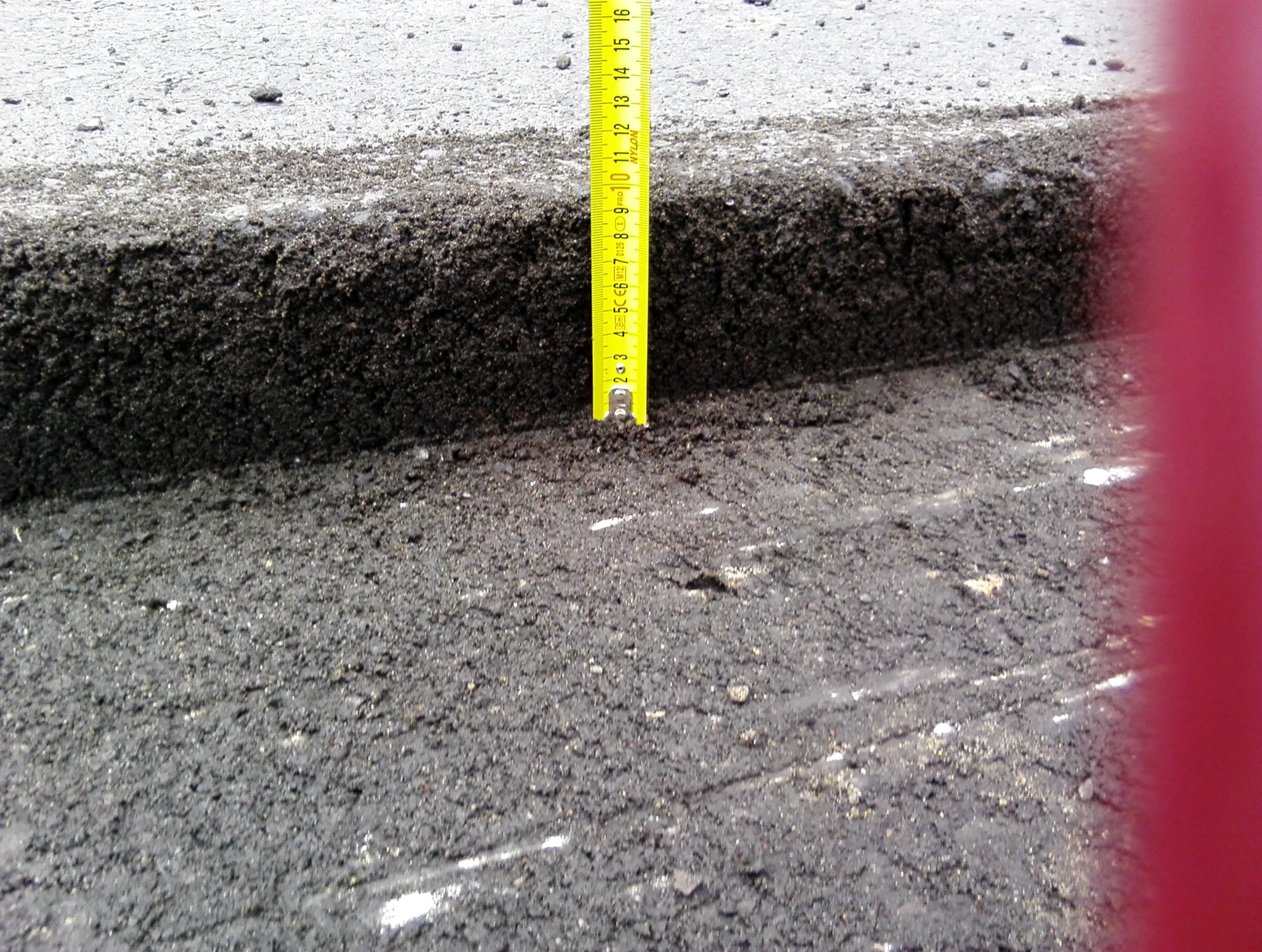 Pomiar grubości warstwy asfaltu żółtą miarką zwijaną na krawędzi nowej nawierzchni drogowej.