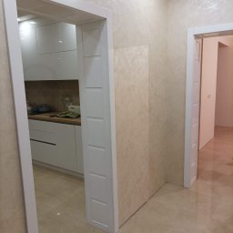 Krzysztof Lis - Beżowe wnętrze po odświeżeniu: ściana z dekoracyjną fakturą, białe drzwi wewnętrzne z frezowanymi panelami, widok na białą kuchnię w głębi.