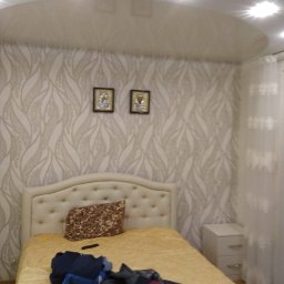 Krzysztof Lis - Sypialnia z łóżkiem z beżowym zagłówkiem, wzorzystą tapetą w odcieniach beżu i brązu oraz dwoma obrazkami na ścianie, oświetlona punktowymi lampami sufitowymi.