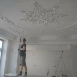 Krzysztof Lis - Pracownik maluje natryskowo sufit z dekoracyjnymi sztukateriami, widoczna drabina i przygotowane ściany w jasnym pomieszczeniu.