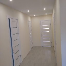 Krzysztof Lis - Korytarz po remoncie z trzema białymi drzwiami z poziomymi przeszkleniami, podłoga wyłożona jasnymi płytkami, oświetlenie punktowe w suficie.