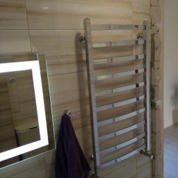 Krzysztof Lis - Chromowany grzejnik łazienkowy drabinkowy zamontowany na beżowych płytkach, obok lustro z podświetleniem LED i fioletowy ręcznik, widoczne niedokończone obrzeże ściany.