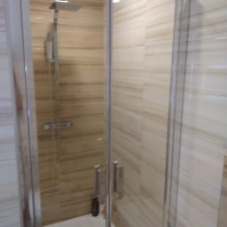 Krzysztof Lis - Kabina prysznicowa z deszczownicą i beżowymi płytkami imitującymi drewno, widoczny odpływ liniowy i siedzisko.