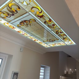 Krzysztof Lis - Sufit z dekoracyjnym, podświetlanym witrażem w odcieniach beżu, żółci i czerwieni, widok z dołu, w tle fragment klatki schodowej z oświetleniem punktowym.