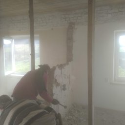 Krzysztof Lis - Dwóch pracowników w zakurzonym pomieszczeniu usuwa starą tapetę ze ściany, przygotowując ją do dalszych prac wykończeniowych.