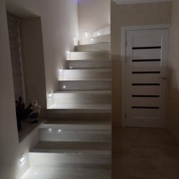 Krzysztof Lis - Nowoczesne schody z podświetleniem LED w ścianie i stopnicach, obok białe drzwi z poziomymi czarnymi paskami, na górze mała choinka.