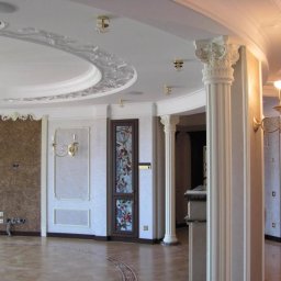 Krzysztof Lis - Eleganckie wnętrze z dekoracyjnymi kolumnami, sztukaterią sufitową i parkietem w odcieniach beżu i brązu, oświetlone złotymi kinkietami i lampami sufitowymi.
