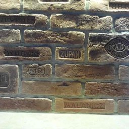 Krzysztof Lis - Dekoracyjna ściana z cegieł z wytłoczonymi napisami i logotypami różnych firm, imitująca starą cegłę.