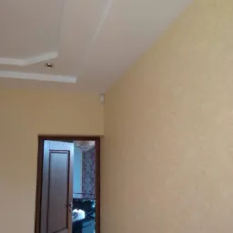 Ściana w jasnym odcieniu beżu z delikatną fakturą, widoczna część sufitu z oświetleniem punktowym i fragment otwartych, ciemnych drzwi z widokiem na wnętrze pokoju z żyrandolem i sofą.