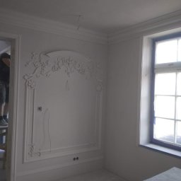 Krzysztof Lis - Białe ściany z dekoracyjnymi sztukateriami, widoczne gniazdka elektryczne i wystające kable, okno z niebieską ramą, fragment drabiny malarskiej z pracownikiem w tle.