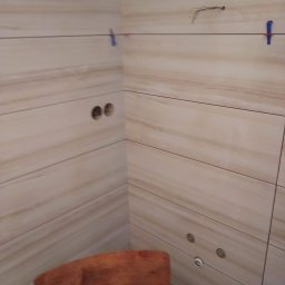 Krzysztof Lis - Ściana w trakcie układania płytek w łazience, widoczne otwory na gniazdka i rury, poziomica i plastikowe krzyżyki do regulacji odstępów.