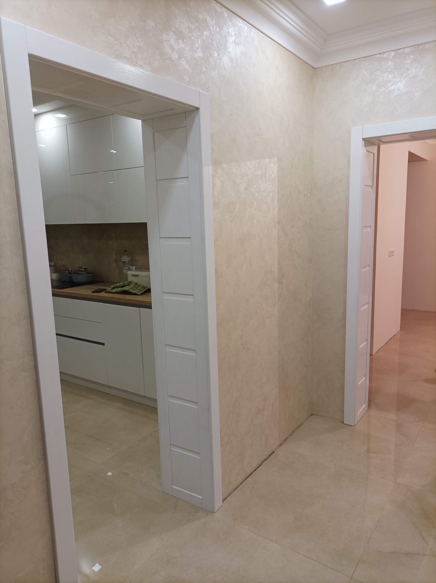 Beżowe wnętrze po odświeżeniu: ściana z dekoracyjną fakturą, białe drzwi wewnętrzne z frezowanymi panelami, widok na białą kuchnię w głębi.