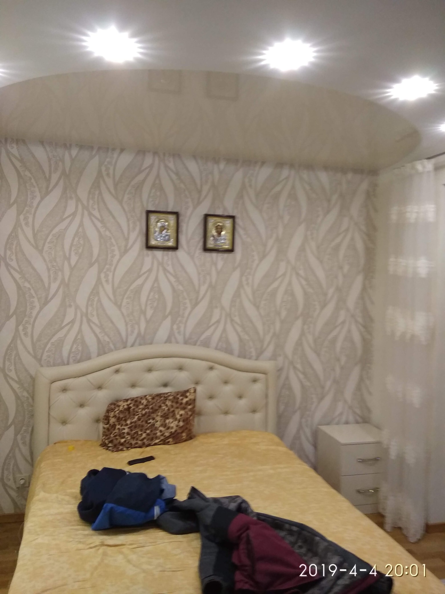 Sypialnia z łóżkiem z beżowym zagłówkiem, wzorzystą tapetą w odcieniach beżu i brązu oraz dwoma obrazkami na ścianie, oświetlona punktowymi lampami sufitowymi.