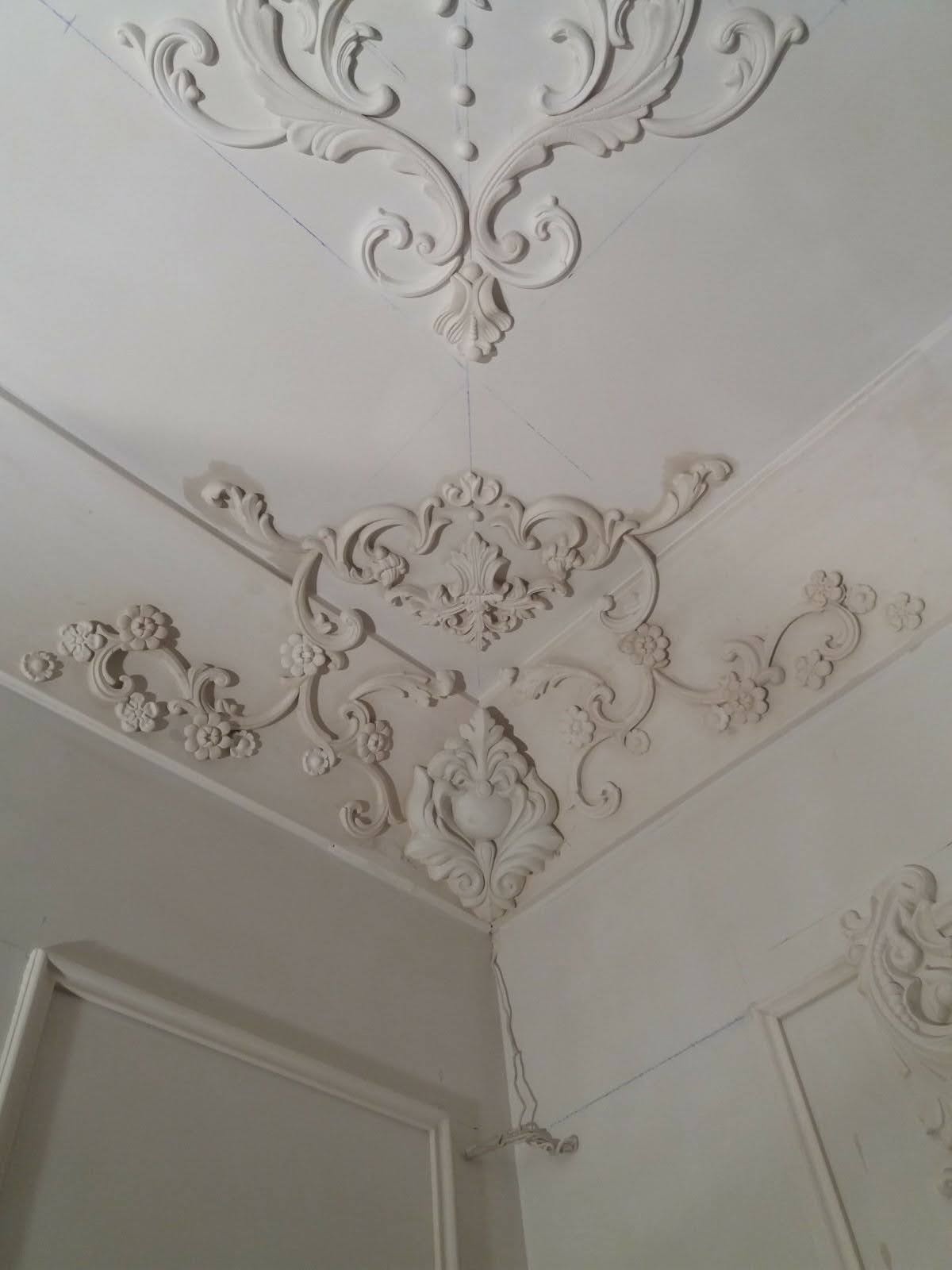 Dekoracyjne sztukaterie narożne na suficie i ścianach, z delikatnymi śladami ołówka wyznaczającego linie, w białym kolorze.