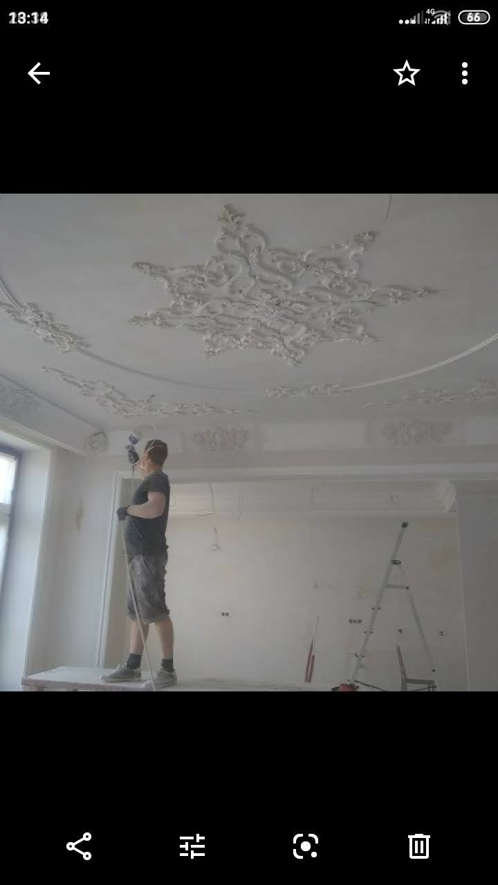 Pracownik maluje natryskowo sufit z dekoracyjnymi sztukateriami, widoczna drabina i przygotowane ściany w jasnym pomieszczeniu.