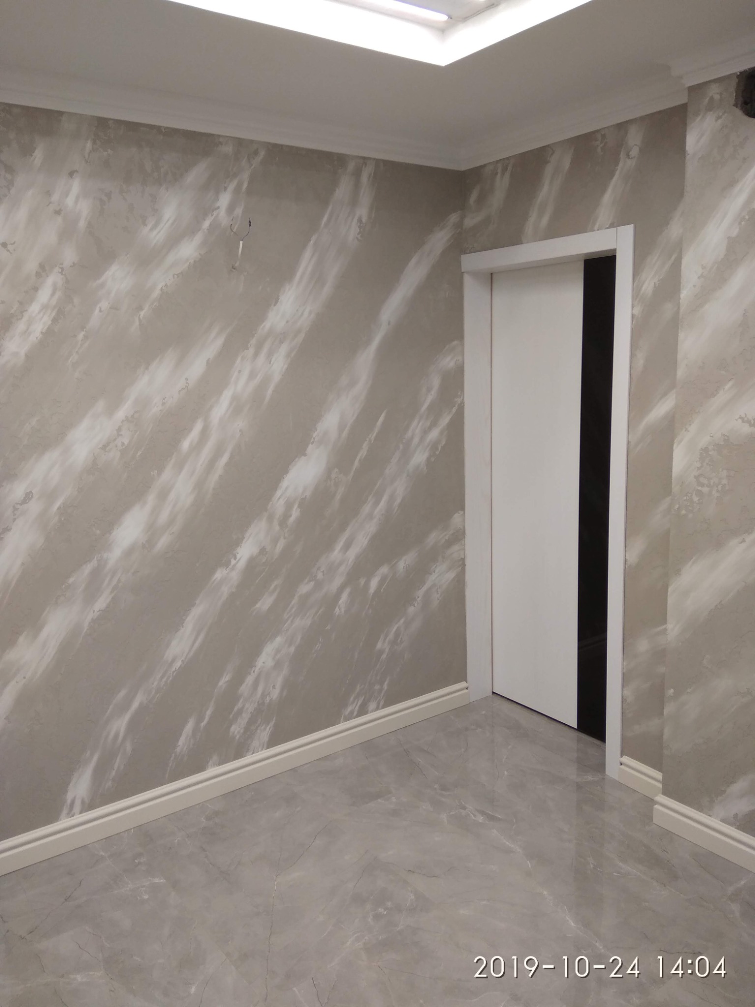 Wykończone wnętrze z dekoracyjną ścianą w odcieniach szarości z efektem marmuru, białe listwy przypodłogowe i sufitowe oraz szara podłoga imitująca marmur, białe drzwi wewnętrzne.