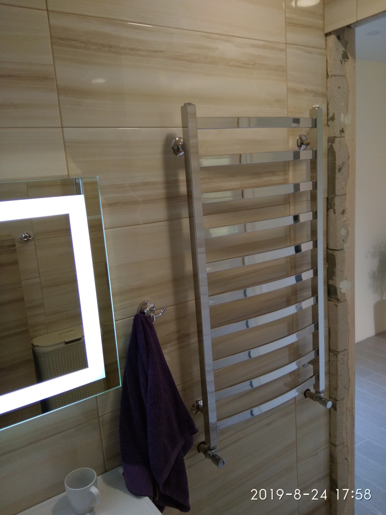 Chromowany grzejnik łazienkowy drabinkowy zamontowany na beżowych płytkach, obok lustro z podświetleniem LED i fioletowy ręcznik, widoczne niedokończone obrzeże ściany.