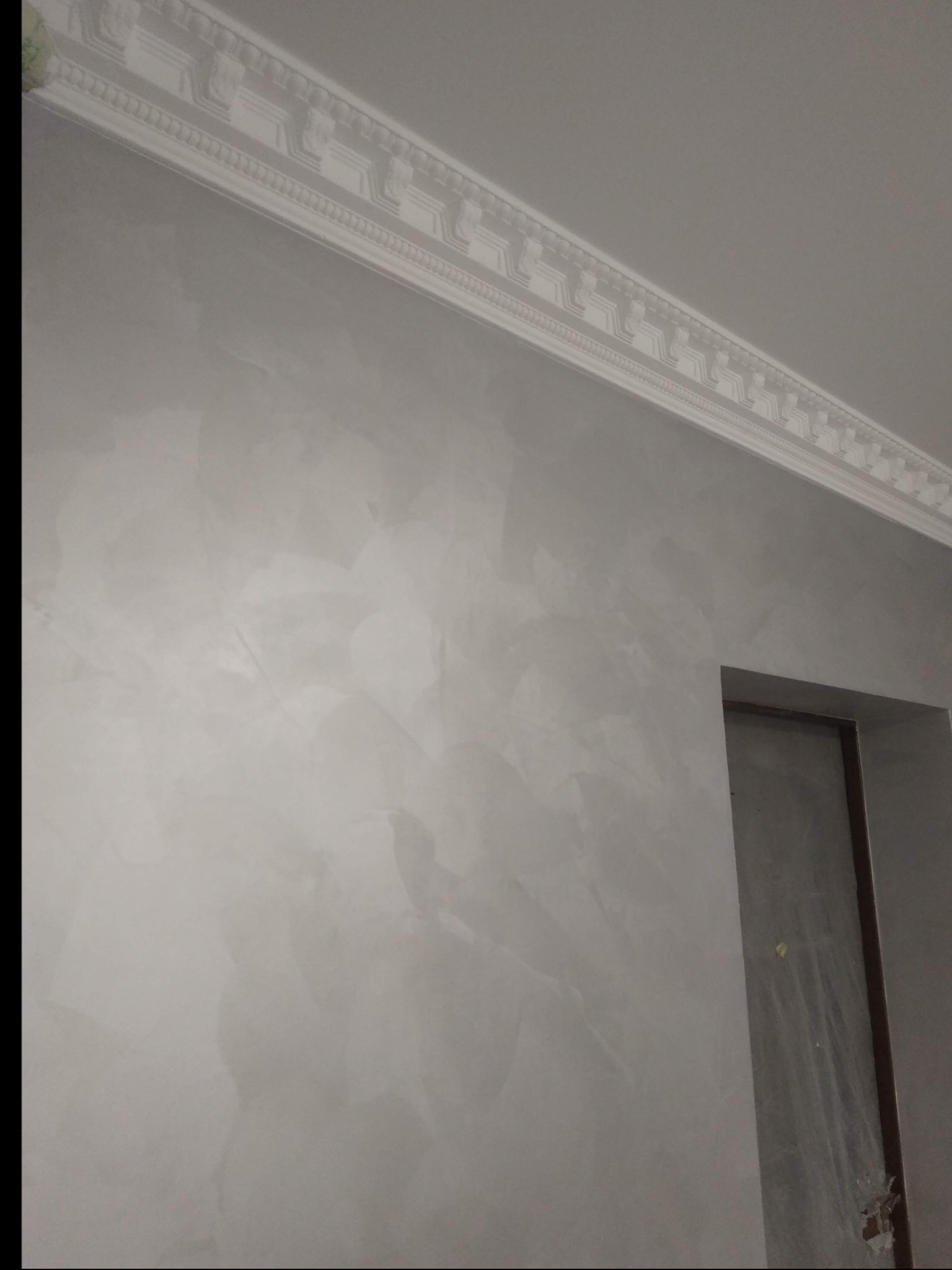 Ściana pokryta dekoracyjną szpachlą w odcieniach szarości z widoczną sztukaterią pod sufitem i ościeżnicą drzwiową.