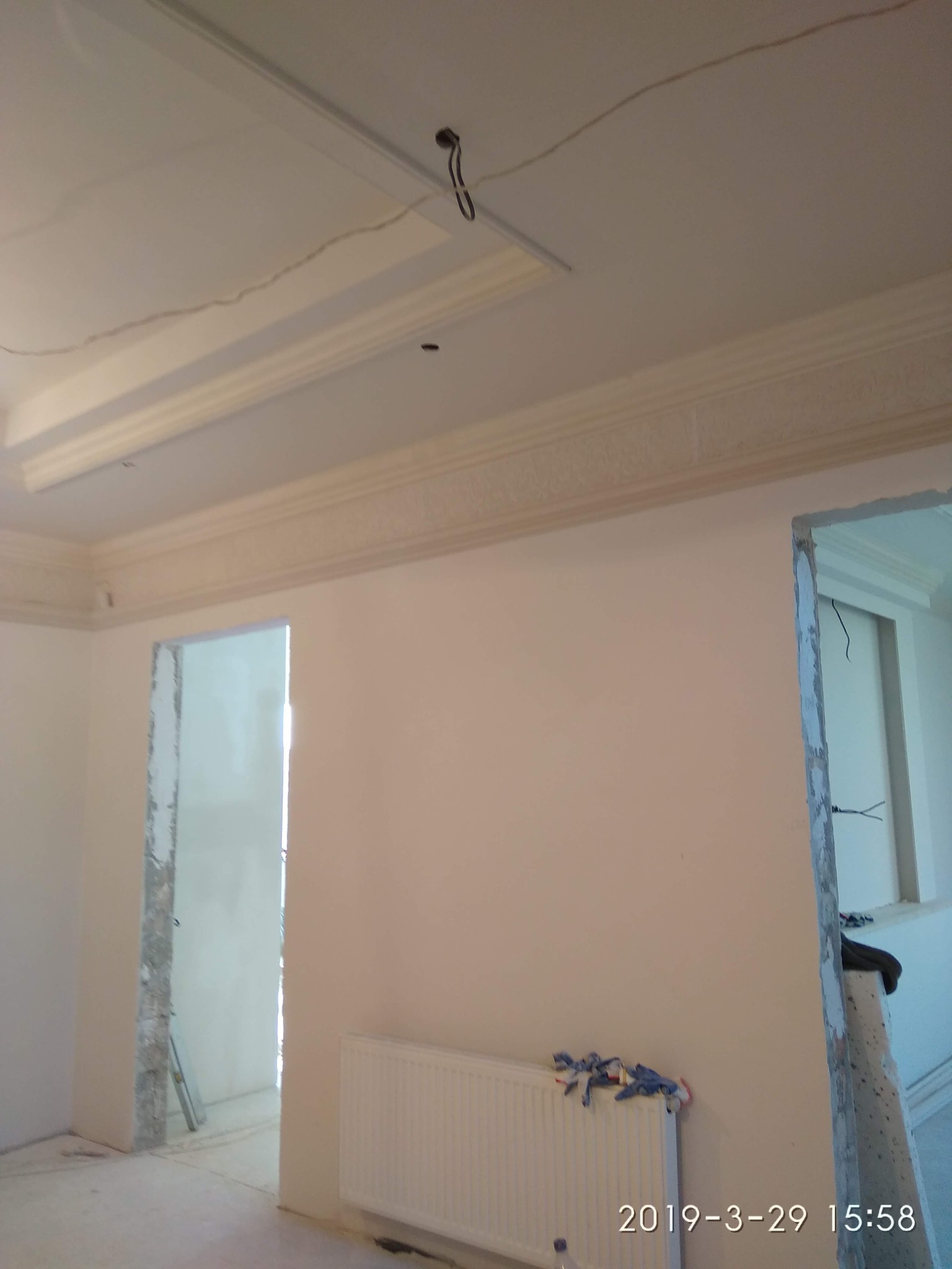 Pokój w trakcie remontu: świeżo pomalowane ściany na kremowo, ozdobny gzyms pod sufitem, widoczne kable i otwór na oświetlenie, grzejnik z roboczymi rękawicami, surowe ościeżnice drzwi.