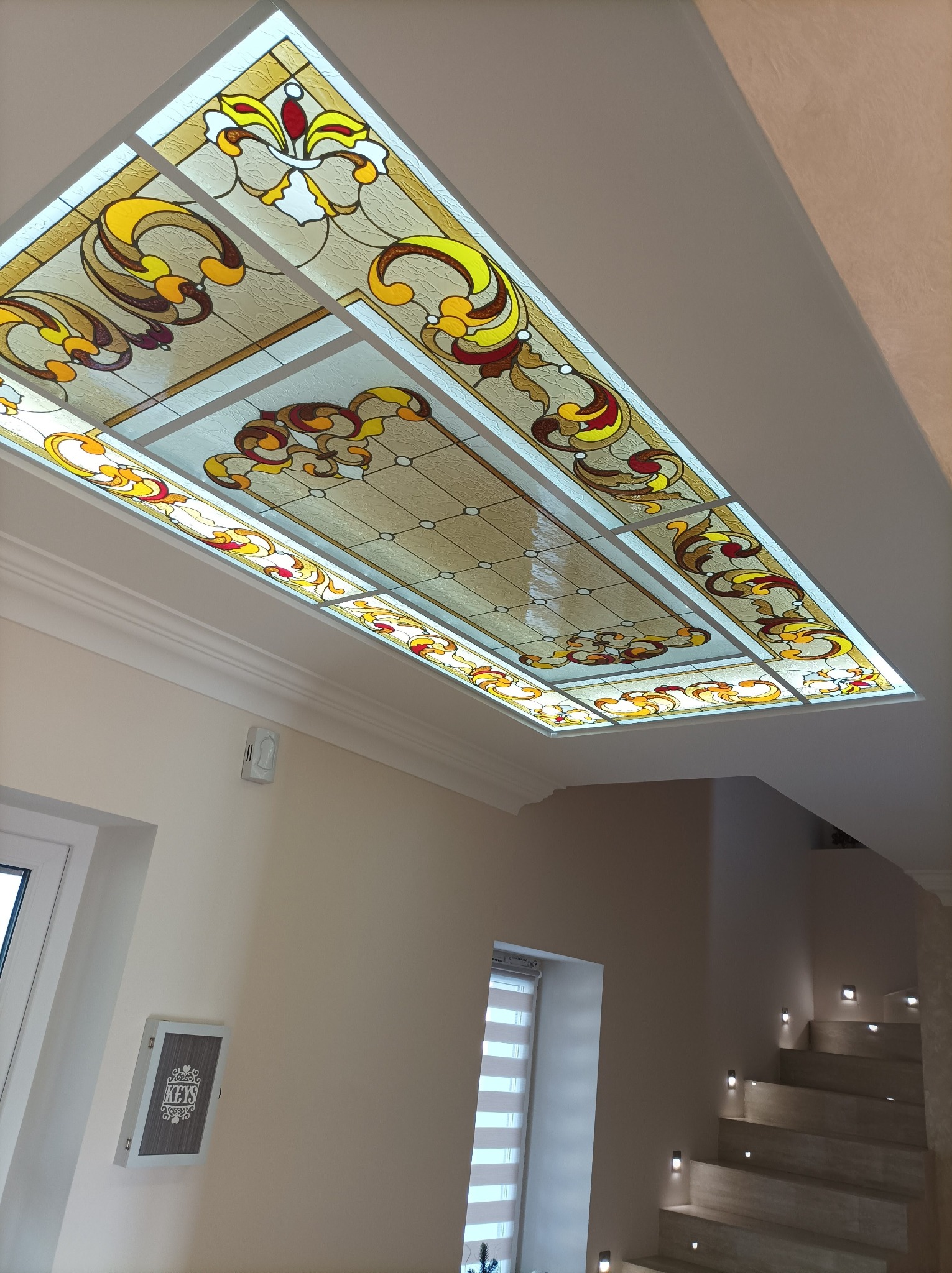 Sufit z dekoracyjnym, podświetlanym witrażem w odcieniach beżu, żółci i czerwieni, widok z dołu, w tle fragment klatki schodowej z oświetleniem punktowym.
