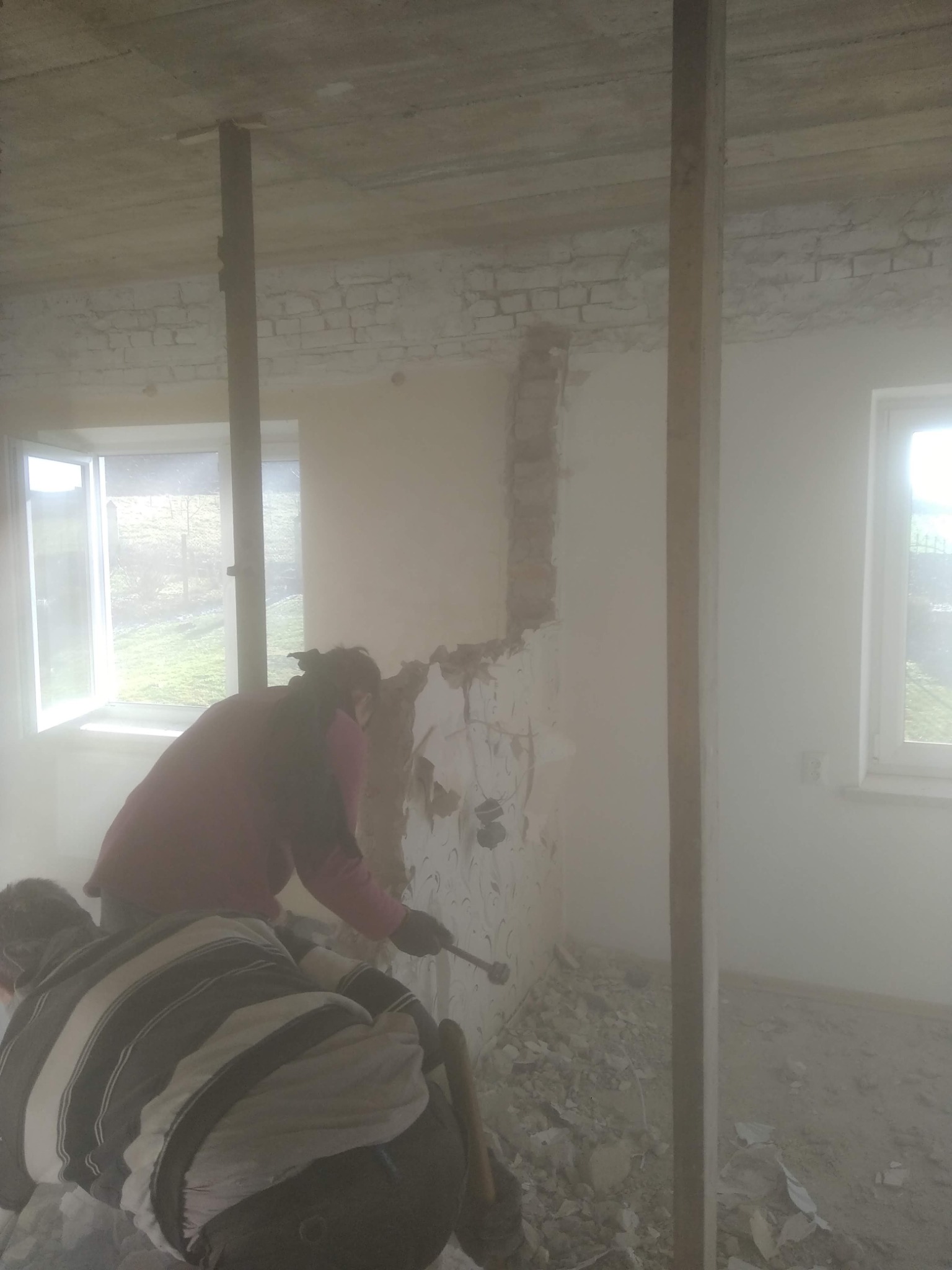 Dwóch pracowników w zakurzonym pomieszczeniu usuwa starą tapetę ze ściany, przygotowując ją do dalszych prac wykończeniowych.