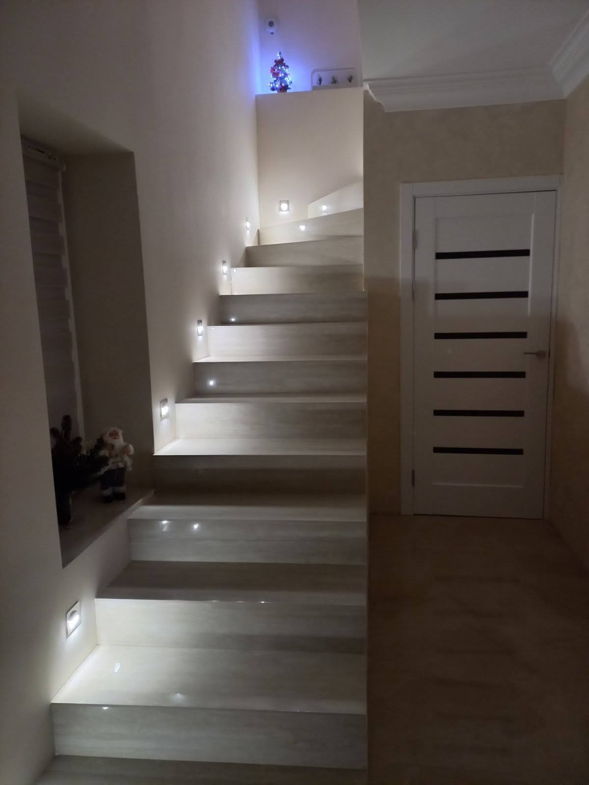 Nowoczesne schody z podświetleniem LED w ścianie i stopnicach, obok białe drzwi z poziomymi czarnymi paskami, na górze mała choinka.