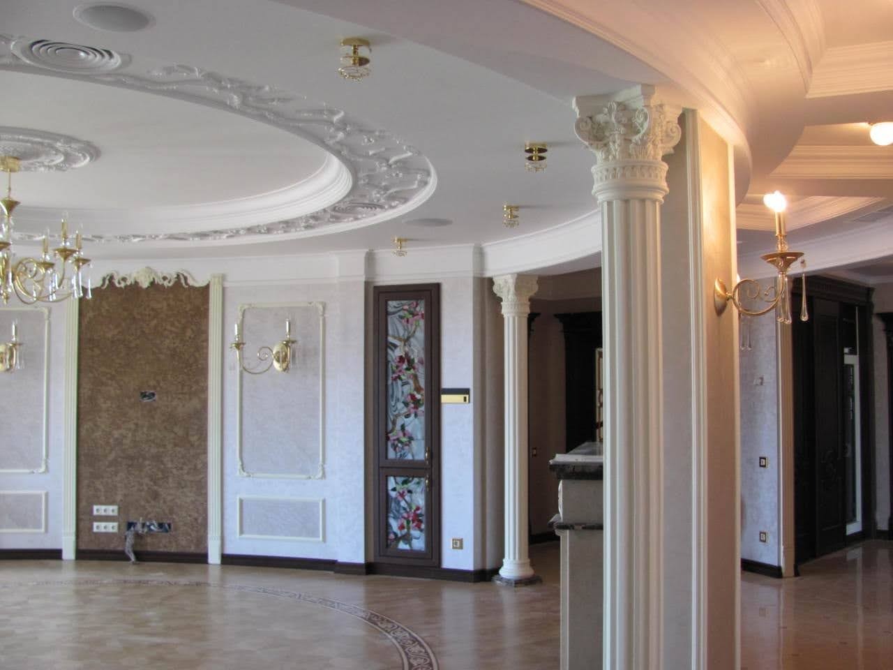 Eleganckie wnętrze z dekoracyjnymi kolumnami, sztukaterią sufitową i parkietem w odcieniach beżu i brązu, oświetlone złotymi kinkietami i lampami sufitowymi.