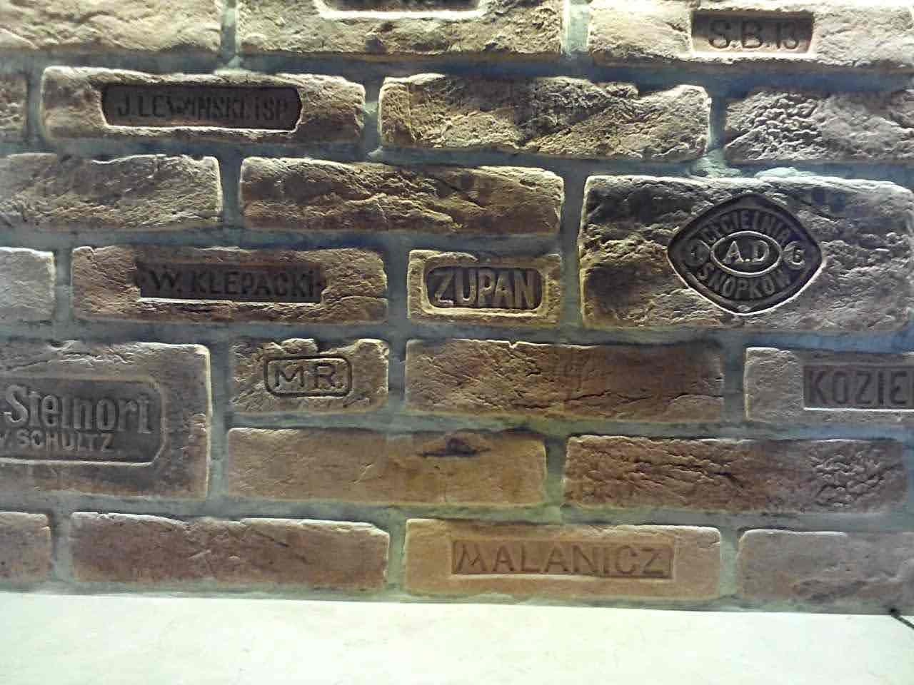 Dekoracyjna ściana z cegieł z wytłoczonymi napisami i logotypami różnych firm, imitująca starą cegłę.