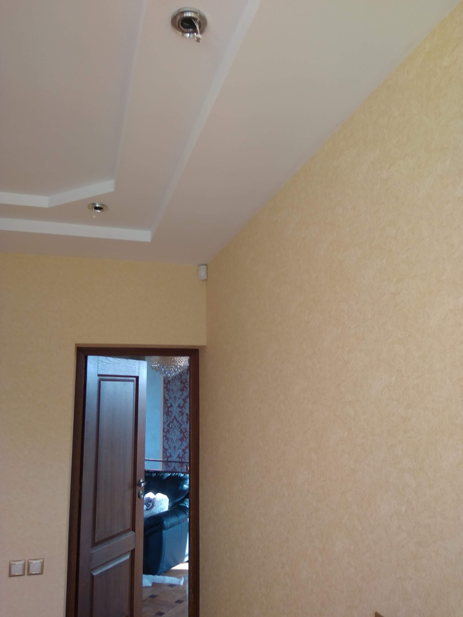 Ściana w jasnym odcieniu beżu z delikatną fakturą, widoczna część sufitu z oświetleniem punktowym i fragment otwartych, ciemnych drzwi z widokiem na wnętrze pokoju z żyrandolem i sofą.