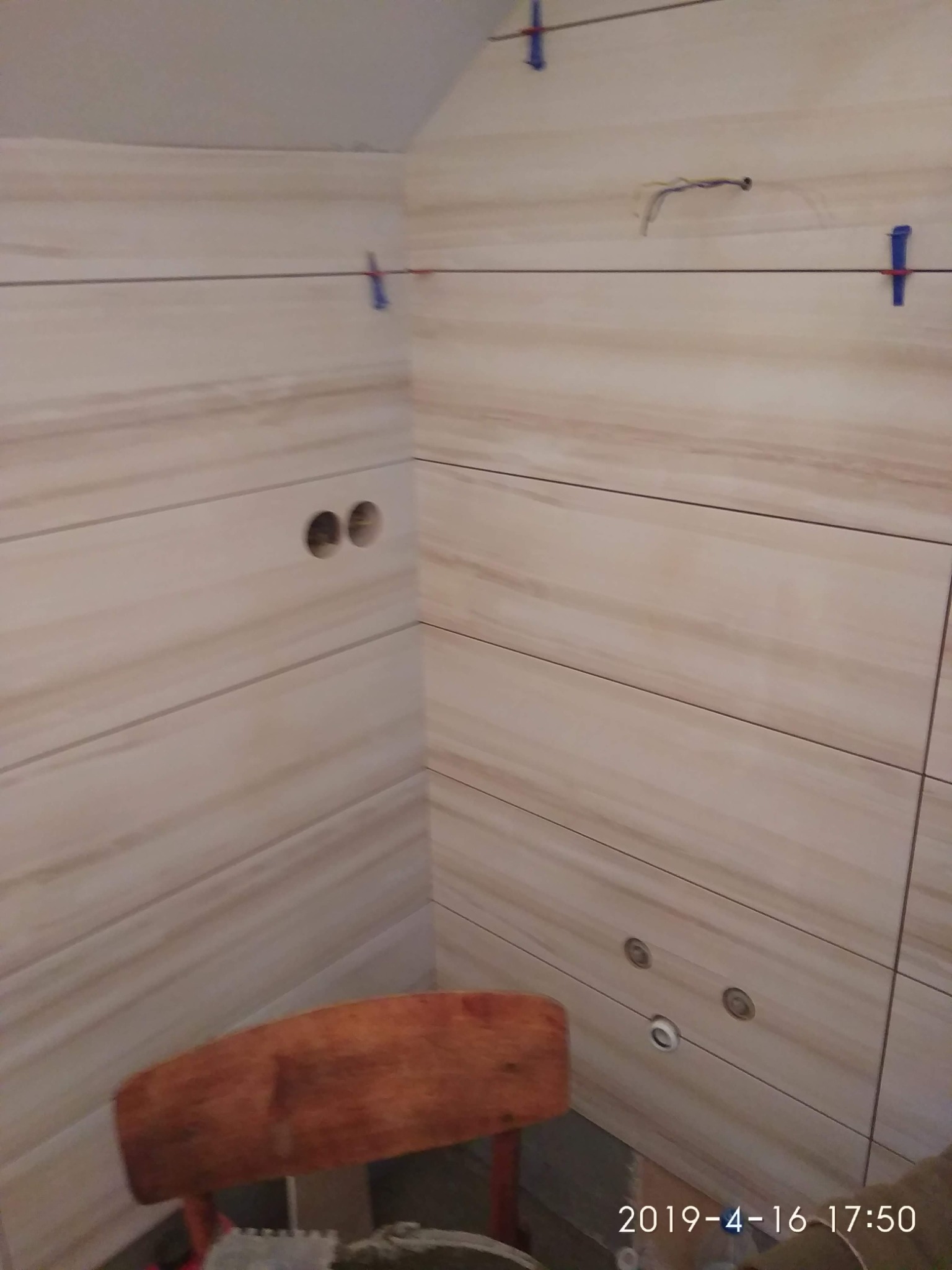 Ściana w trakcie układania płytek w łazience, widoczne otwory na gniazdka i rury, poziomica i plastikowe krzyżyki do regulacji odstępów.