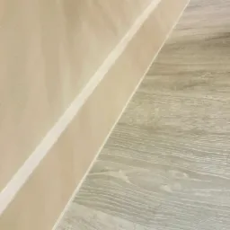 Beżowa ściana z białą listwą przypodłogową sąsiadująca z panelami podłogowymi w odcieniach szarości z widoczną strukturą drewna.