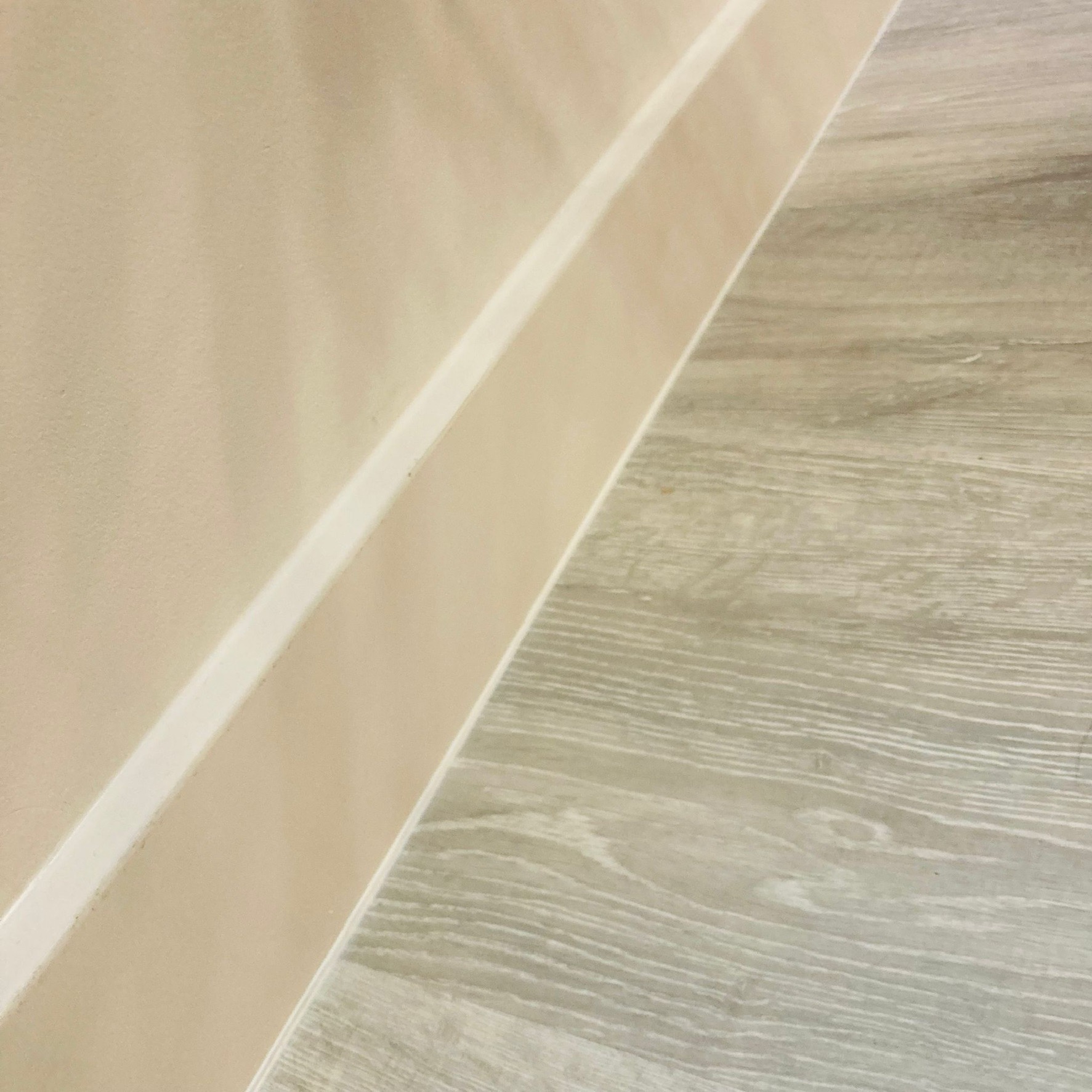 Beżowa ściana z białą listwą przypodłogową sąsiadująca z panelami podłogowymi w odcieniach szarości z widoczną strukturą drewna.