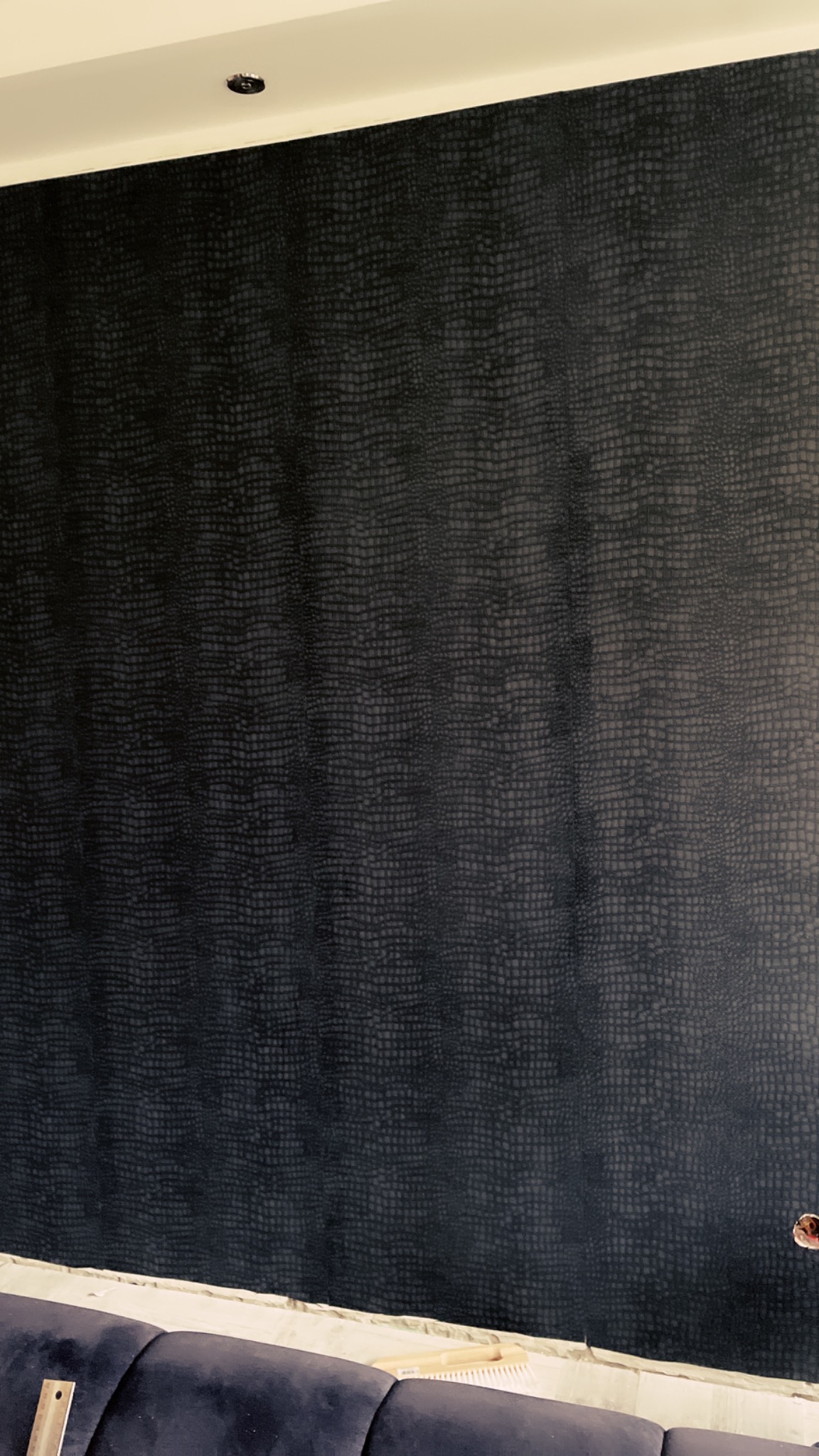 Ściana pokryta ciemną tapetą o wzorze imitującym skórę krokodyla, widoczny fragment sofy i listwa przypodłogowa w trakcie montażu.