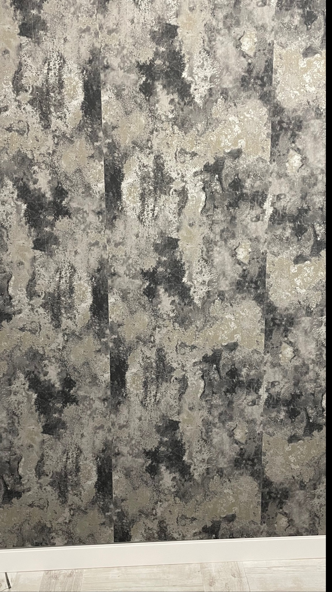 Fragment ściany pokrytej tapetą o wzorze imitującym naturalny kamień w odcieniach szarości i beżu, widoczna biała listwa przypodłogowa i fragment podłogi.