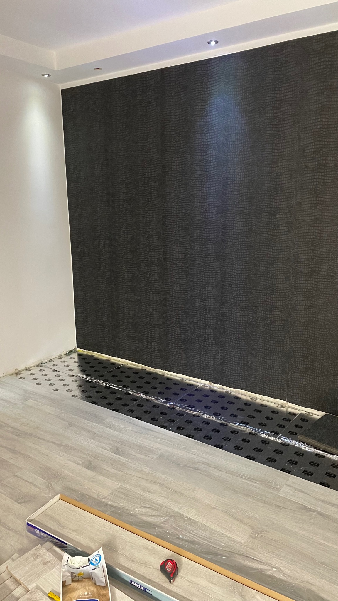 Ściana pokryta tapetą o wzorze skóry węża, podłoga w trakcie układania paneli, widoczne opakowania paneli i miarka.