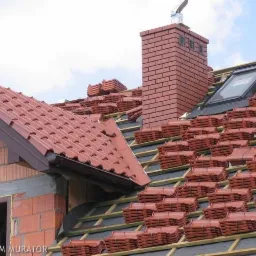 Układanie czerwonej dachówki ceramicznej na dachu z oknem połaciowym i kominem z cegły, widoczny fragment konstrukcji więźby dachowej i przygotowane pakiety dachówek.