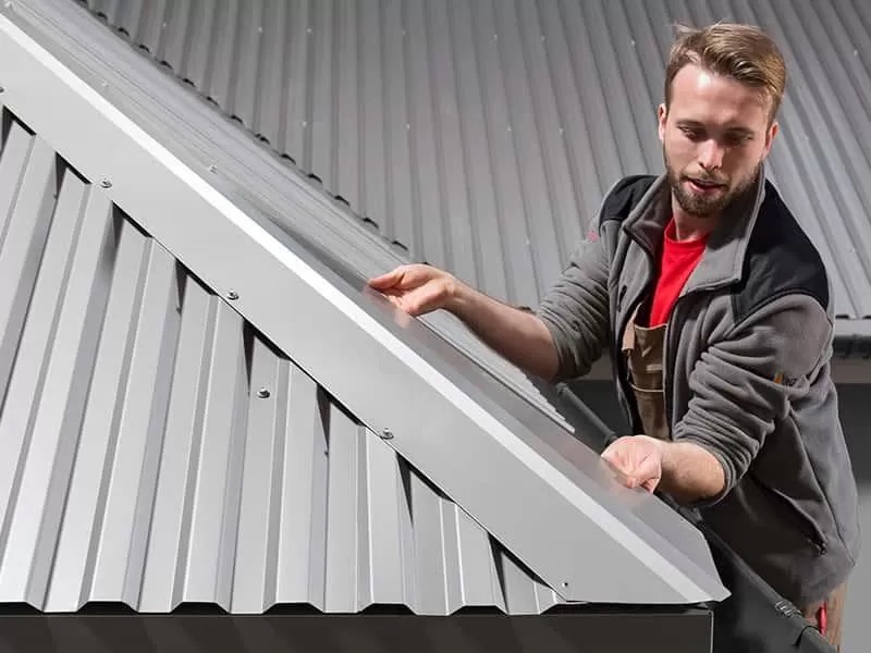 Montaż metalowej listwy wykończeniowej na dachu z blachy trapezowej przez dekarza w szarej bluzie roboczej.