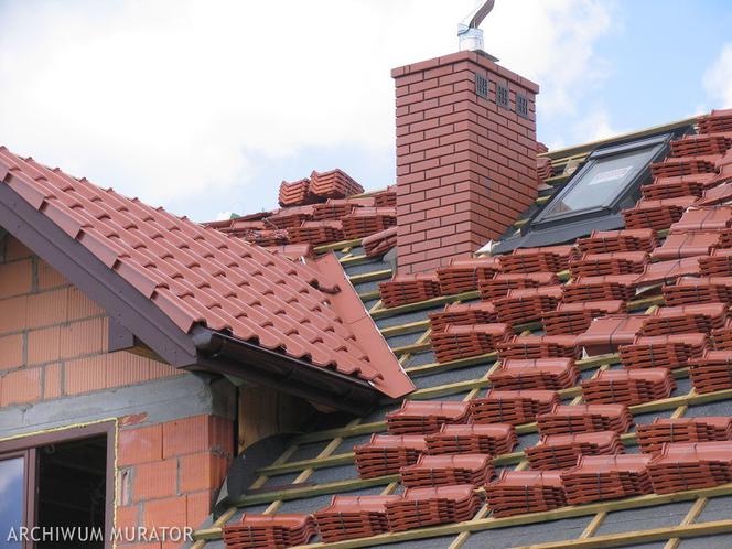 Układanie czerwonej dachówki ceramicznej na dachu z oknem połaciowym i kominem z cegły, widoczny fragment konstrukcji więźby dachowej i przygotowane pakiety dachówek.